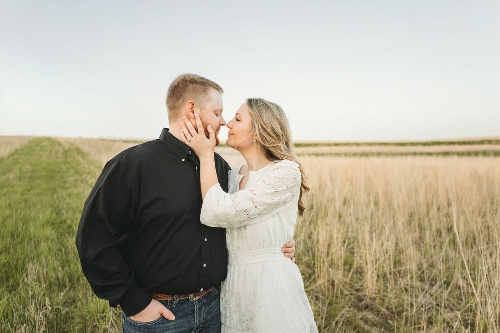 Couple sharing a romantic almost-kiss moment in a golden wheat field, woman in white lace dress and man in black shirt.