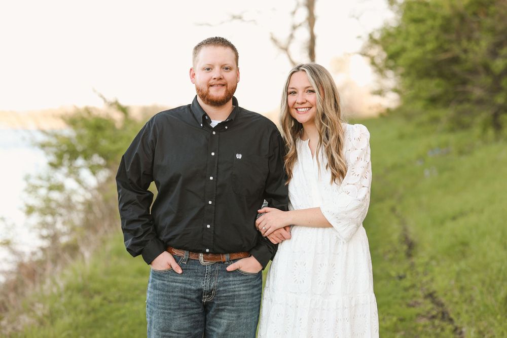 A smiling couple poses outdoors on a green hillside near water, man in black shirt and jeans, woman in white dress.