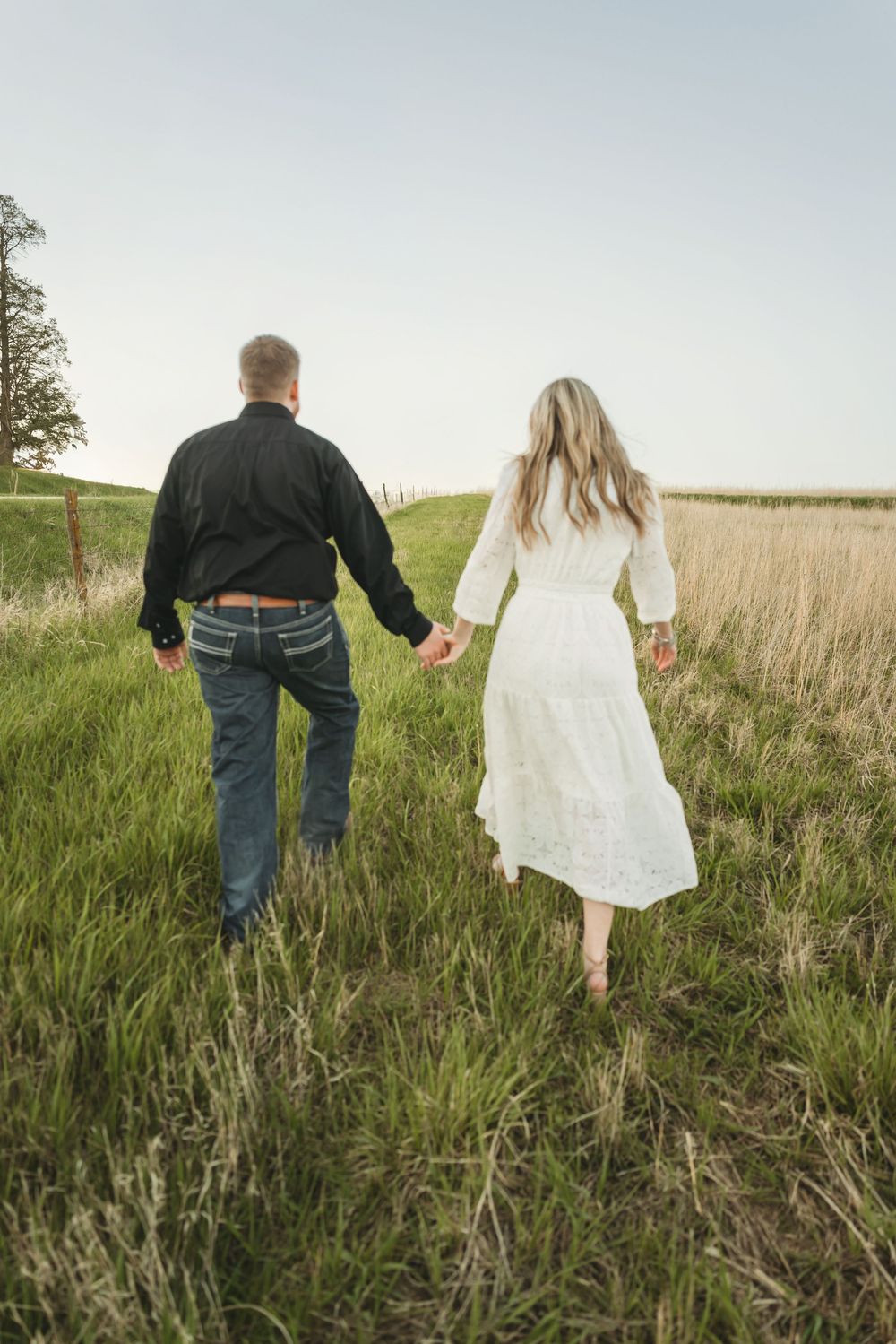Couple holding hands walking through lush green field, woman in white maxi dress and man in black jacket and jeans.