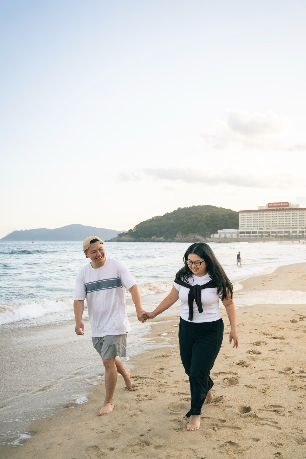 Couple holding hands and walking barefoot on a sandy beach at sunset with a hotel and mountains in the background.