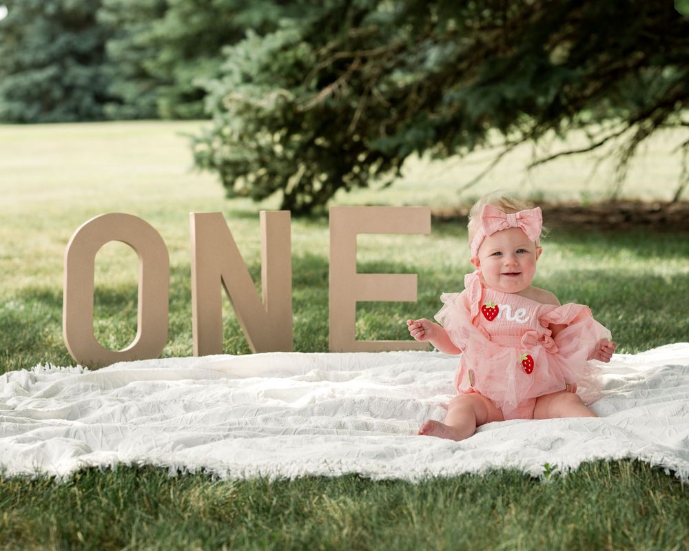 One-year-old girl in a pink dress sitting outdoors during her strawberry shortcake-themed first birthday session.