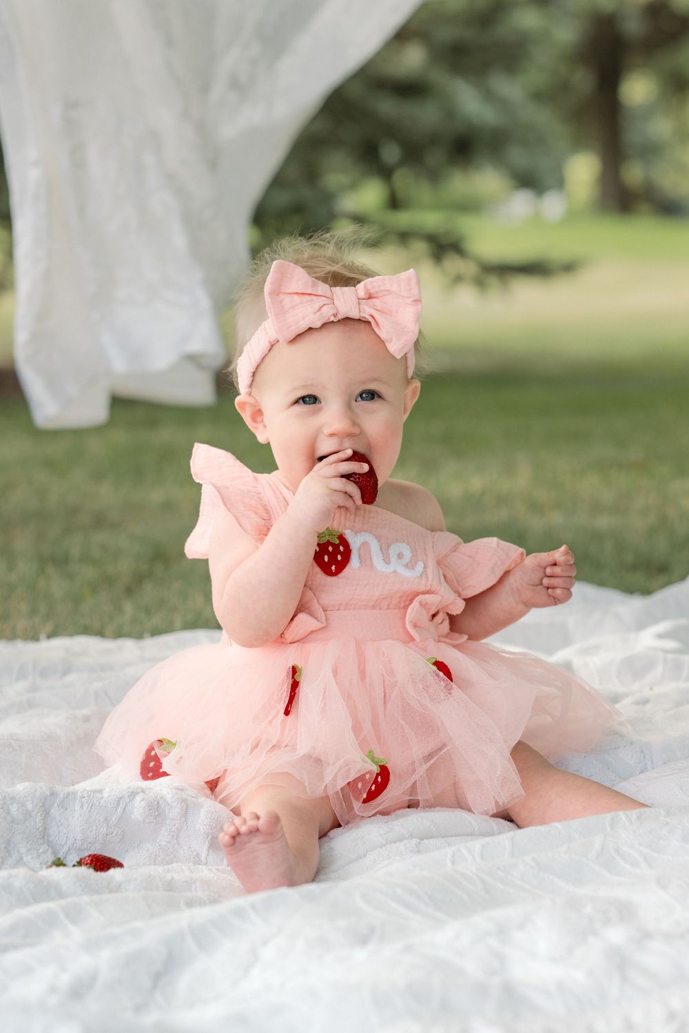 Smiling one-year-old in a pink birthday dress surrounded by strawberries and sweet first birthday decorations.