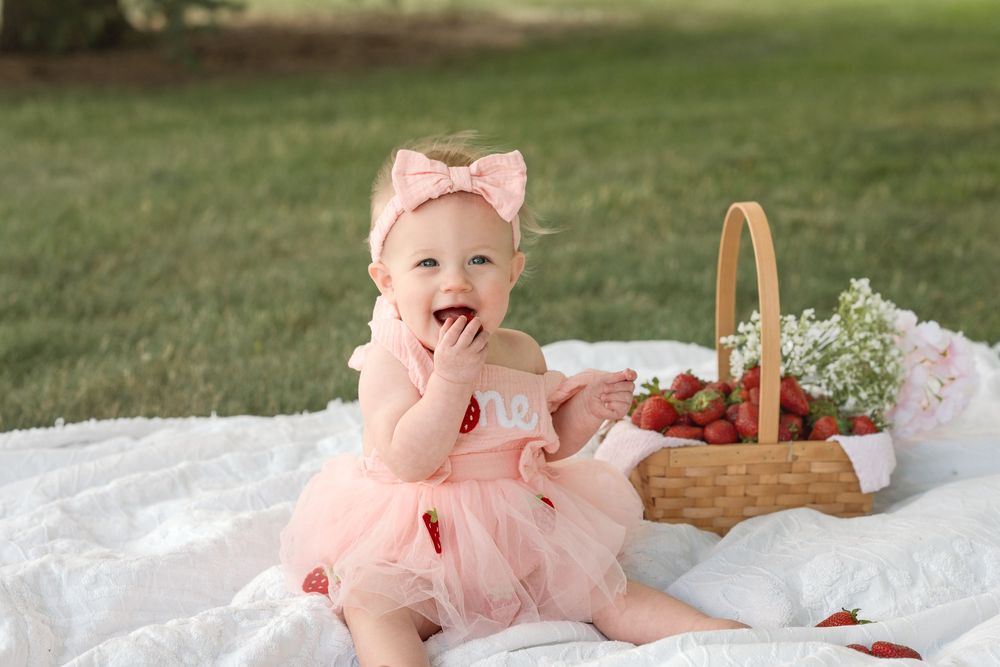 Birthday girl holding a strawberry while wearing a pink dress for her one-year milestone photos.