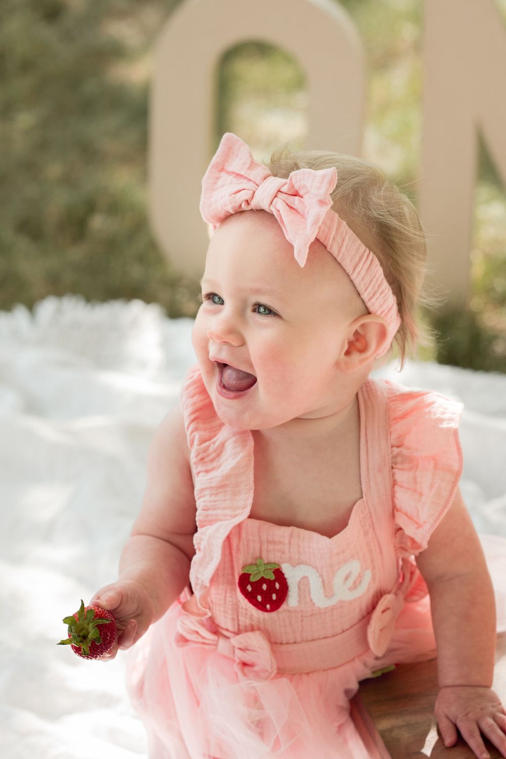 Birthday girl holding a strawberry while wearing a pink dress for her one-year milestone photos.