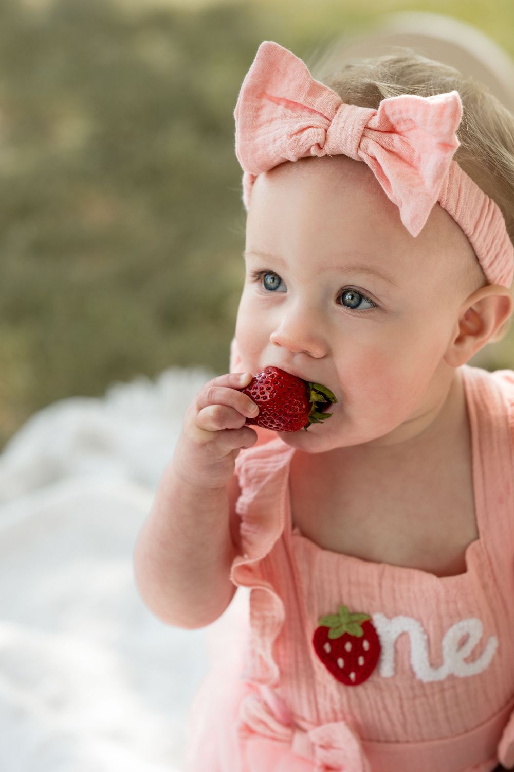 Sweet outdoor portrait of a baby girl celebrating her first birthday with a pink strawberry shortcake theme eating strawberry.