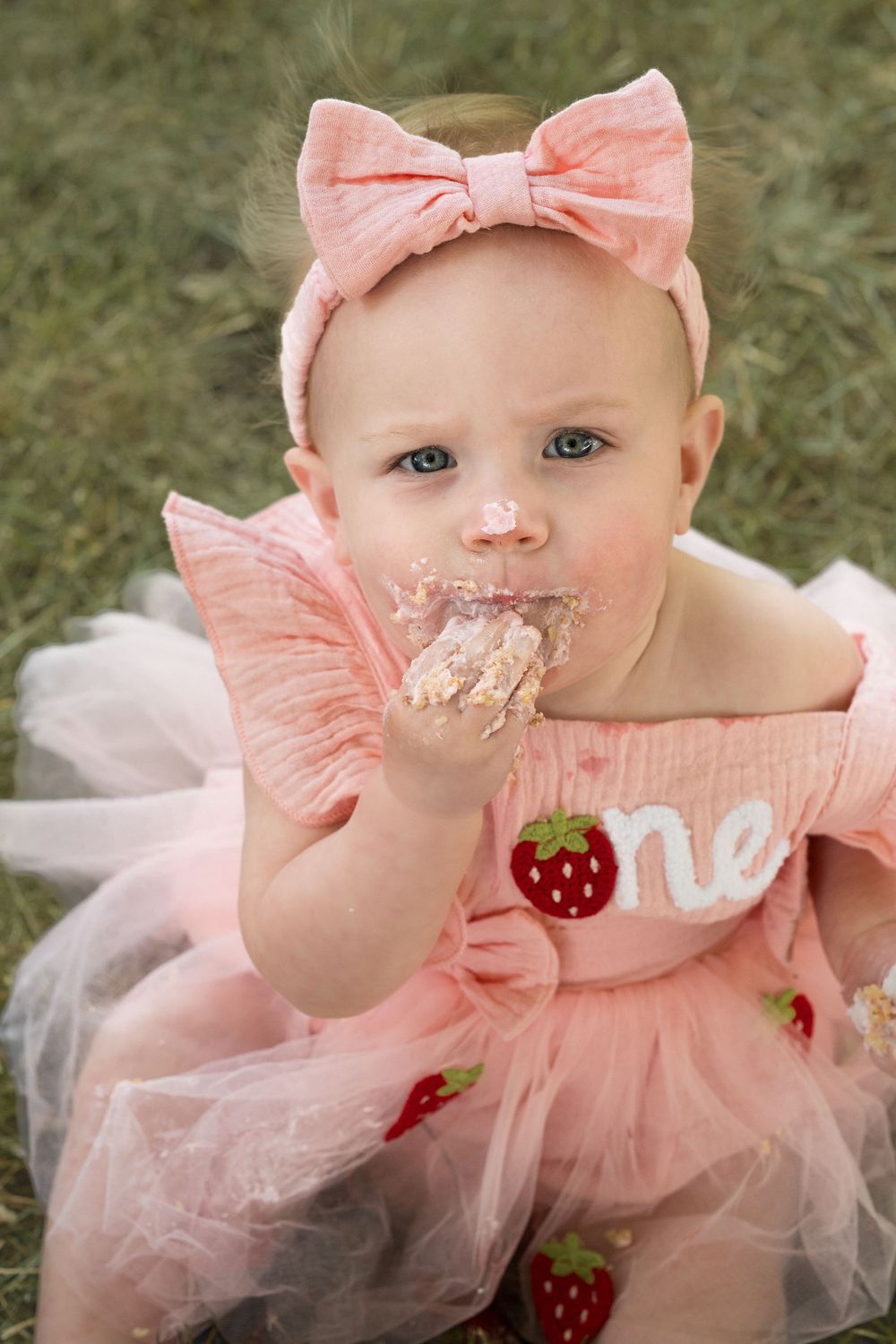 Sweet outdoor portrait of a baby girl celebrating her first birthday with a pink strawberry shortcake theme during cake smash.