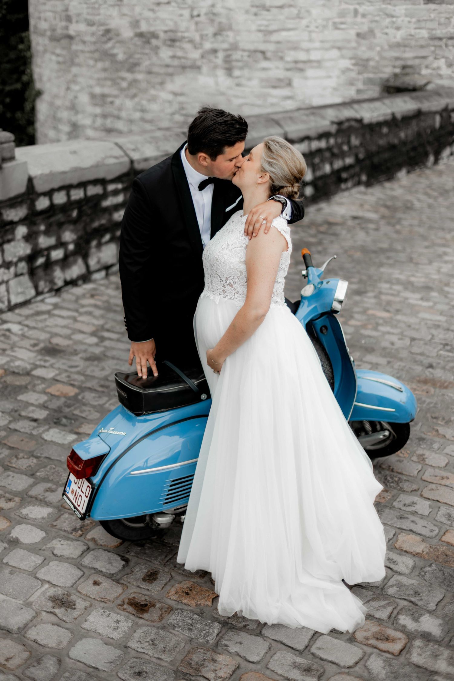 Bride and groom share a romantic kiss beside a vintage blue Vespa scooter on a cobblestone street.