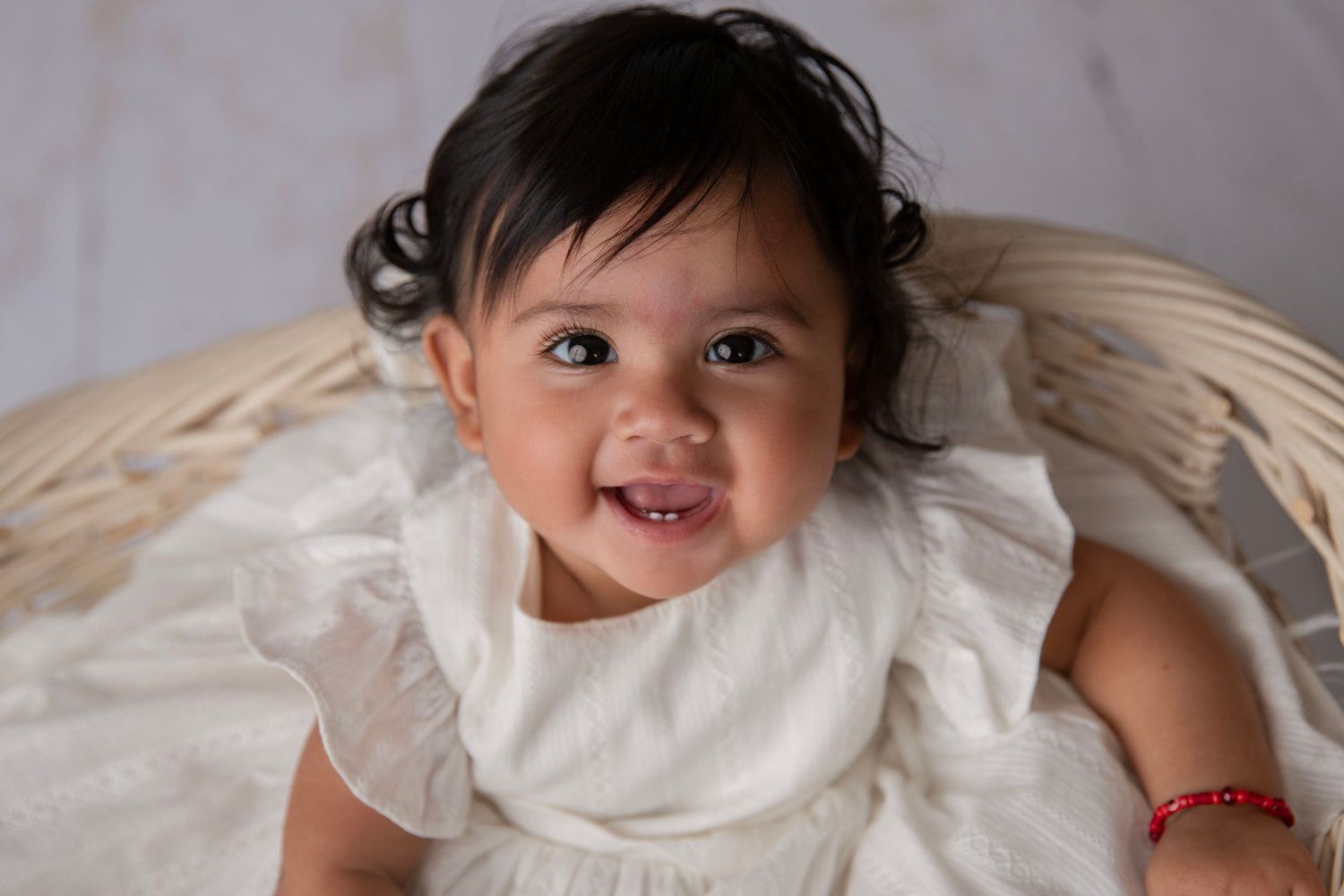 Smiling baby girl in white dress sitting in wicker basket, wearing red bracelet.