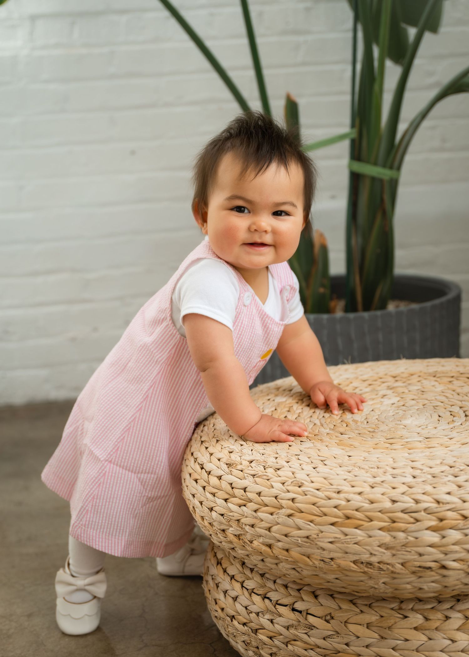A joyful young girl in a pink smocked dress and white shoes sits laughing on a bench indoors.