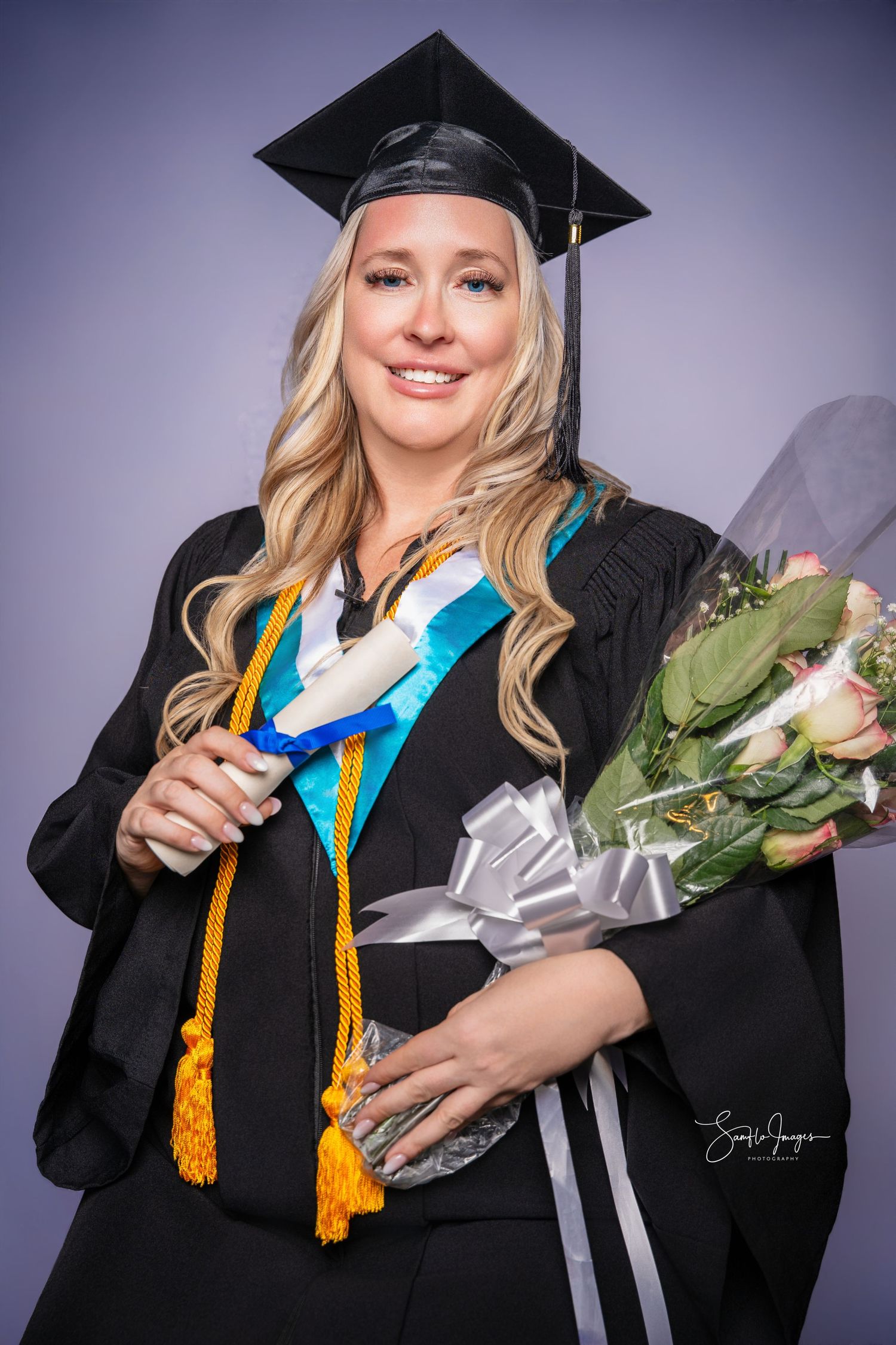 Woman in black graduation gown and cap holding flowers and diploma, wearing gold honor cords and colorful stole.