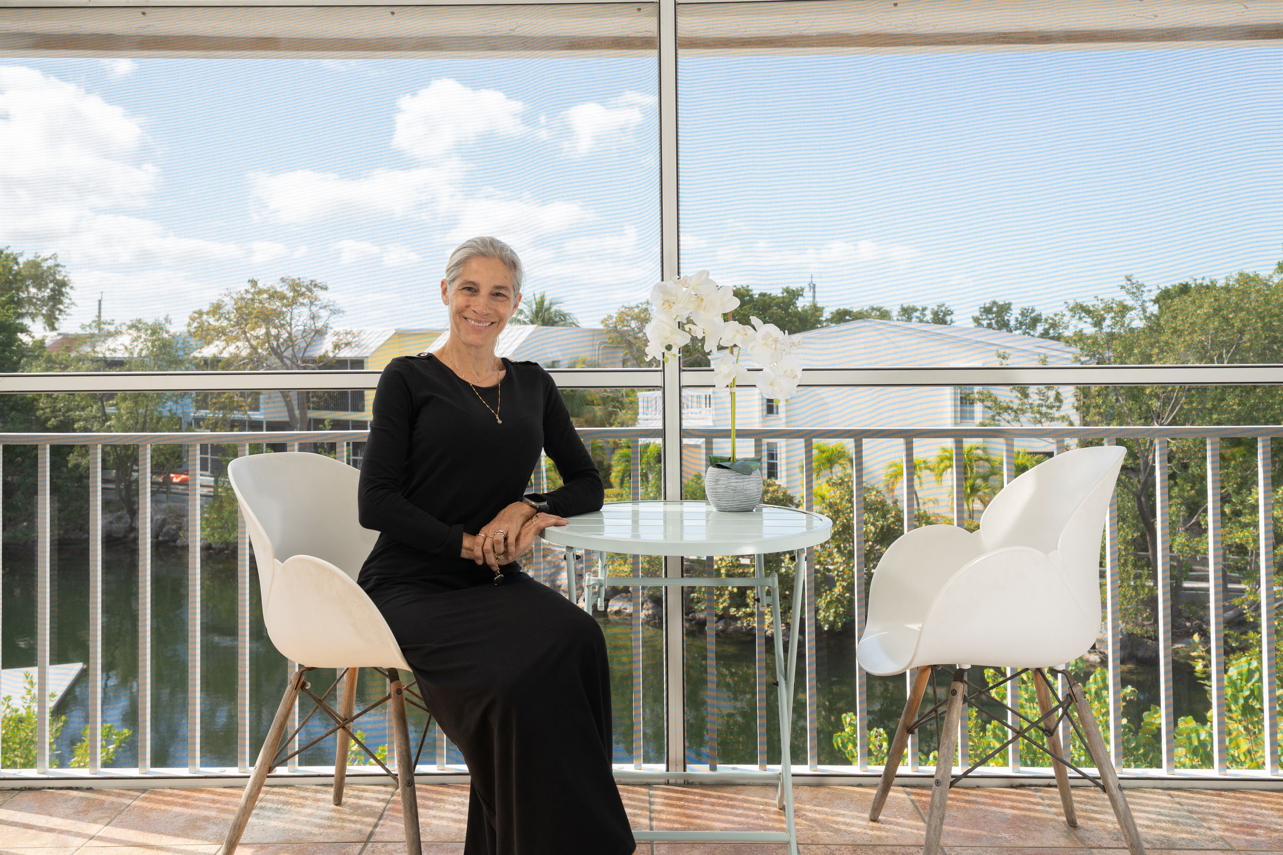 Woman in black dress sitting on modern balcony with white chairs and table, surrounded by lush green trees.