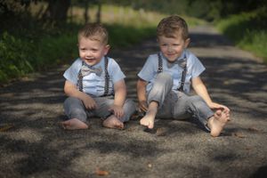 Two blonde toddler twin boys in suspenders and bow ties sitting on a shaded garden path.