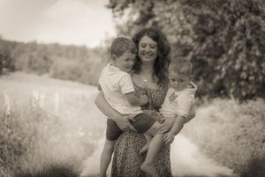 Black and white photo of smiling mother holding two young children on a sunny outdoor path.