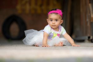 Cute baby girl in white tutu skirt and pink headband crawling on ground outdoors.