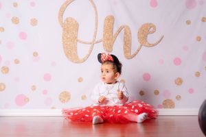 Cute baby girl in red polka dot tutu and Minnie ears poses at One first birthday backdrop.