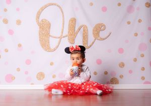 Baby girl in red tutu and Minnie ears holds treat at glittery One first birthday backdrop.