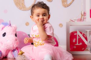 Adorable baby girl in pink tutu dress sitting with unicorn plush toy at first birthday party decoration.