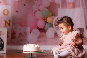 Baby girl in pink dress gazes at white first birthday cake with balloon backdrop behind her.
