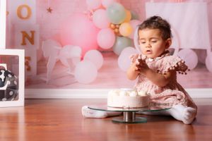 Baby girl in pink dress reaches into first birthday cake during cake smash with balloon decor.