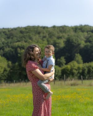 Happy smiling mother in red floral dress holding young son in green flower meadow on sunny day.