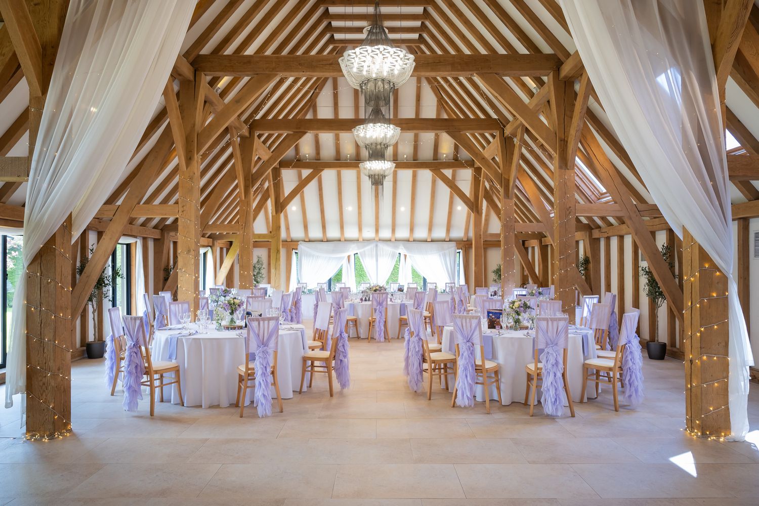 Elegant Old Kent Barn wedding reception with draped white fabric, round tables, lavender chair sashes, and fairy lights.