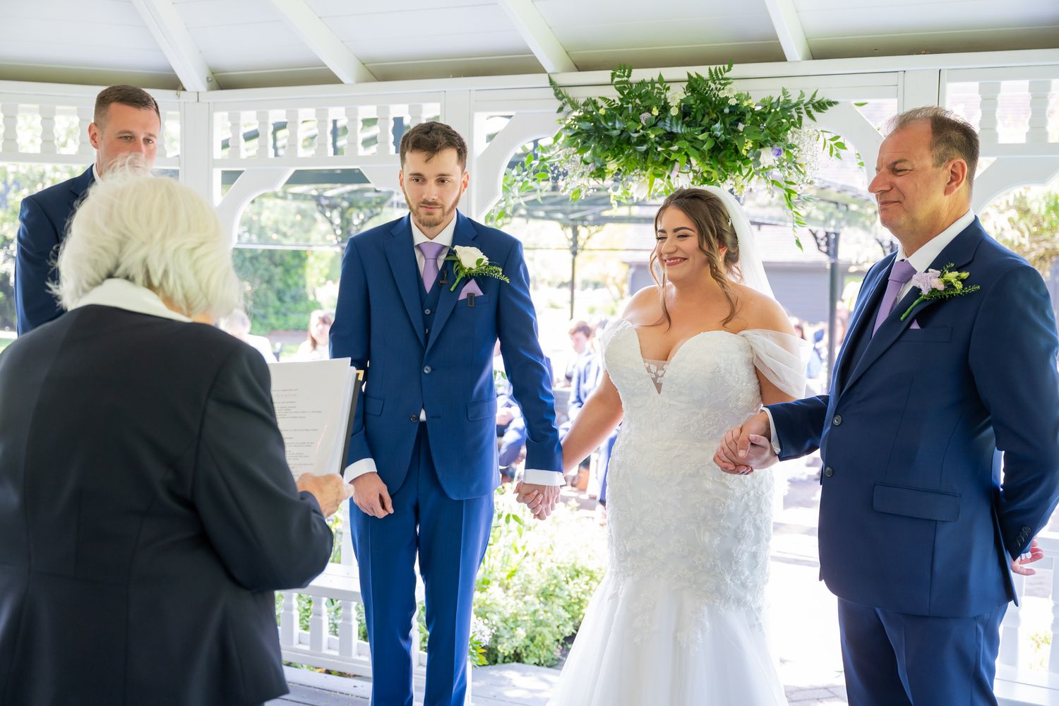 Bride and groom exchanging vows during outdoor wedding ceremony with officiant and father present.