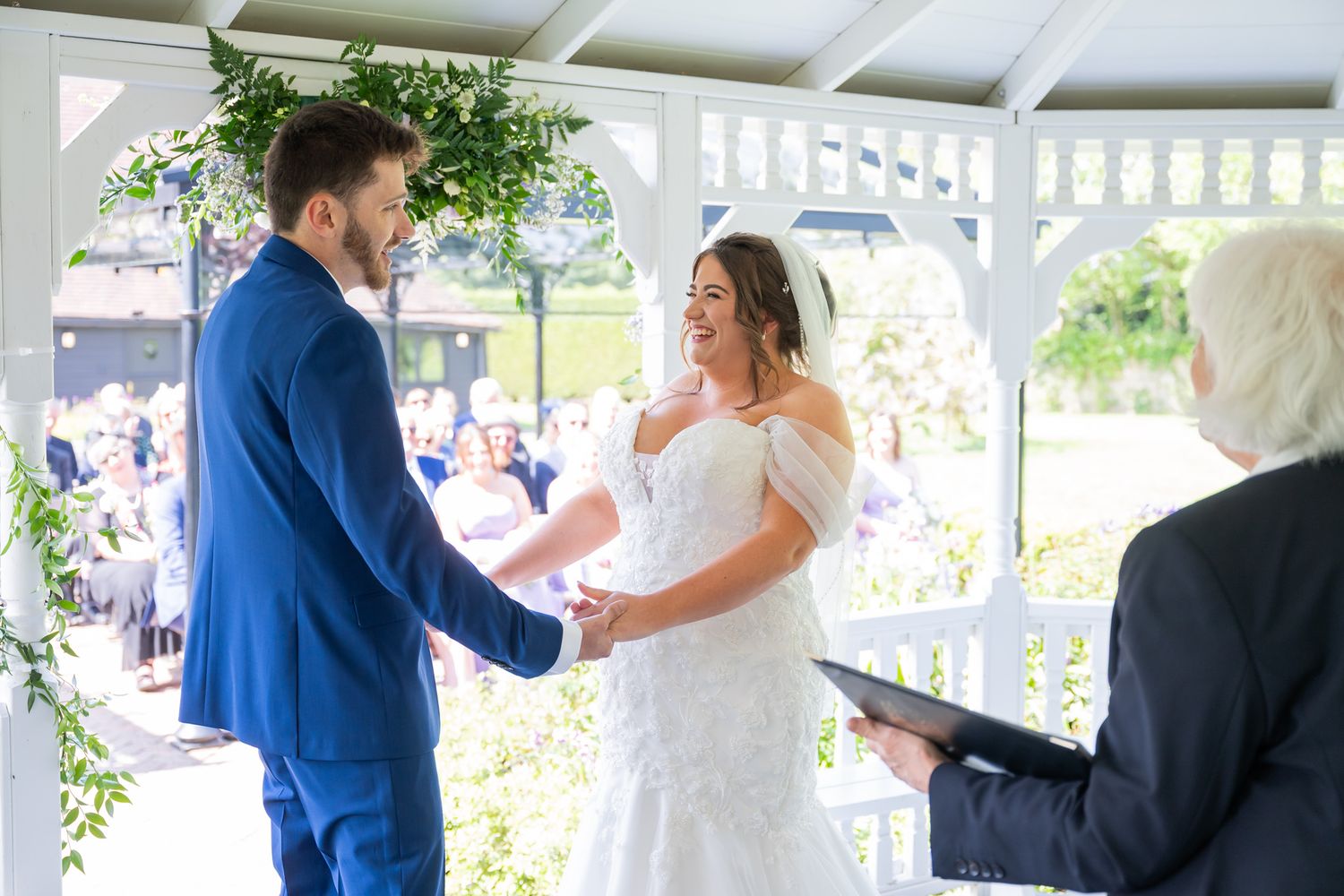 Bride and groom exchange vows at outdoor wedding ceremony in white gazebo adorned with greenery.