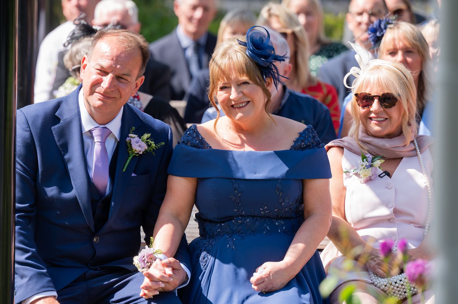 Smiling wedding guests in blue outfits seated at an outdoor wedding ceremony.