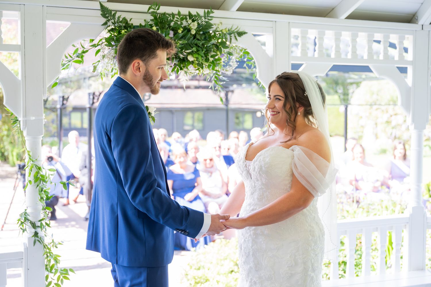 Bride and groom holding hands at outdoor wedding ceremony in white gazebo decorated with greenery.