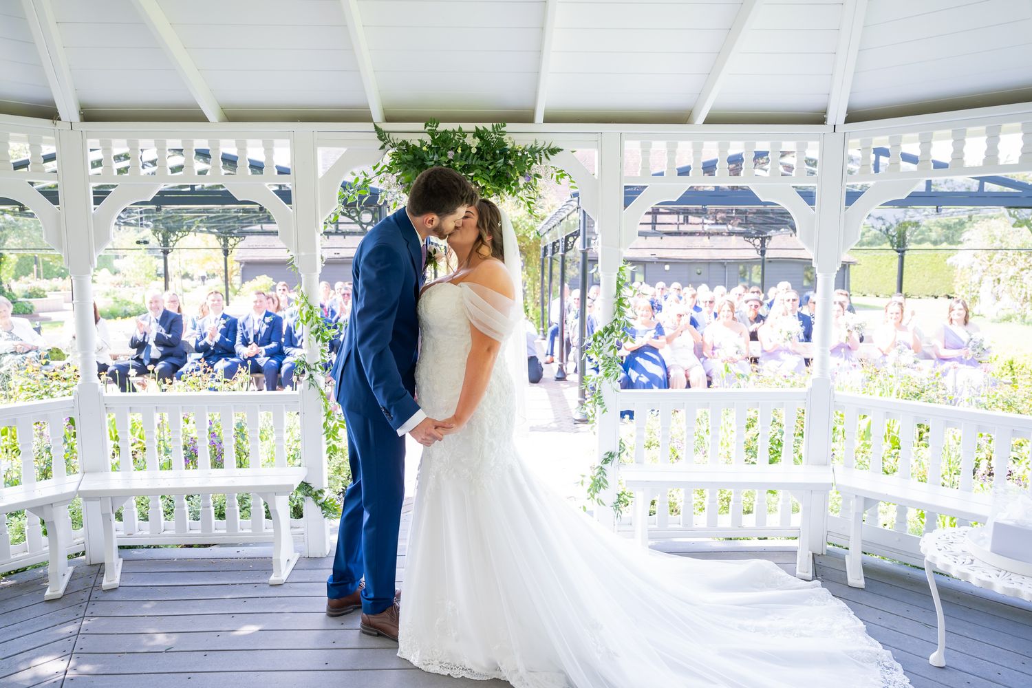 Bride and groom share a romantic moment under a floral arch in a white gazebo during an outdoor wedding ceremony.
