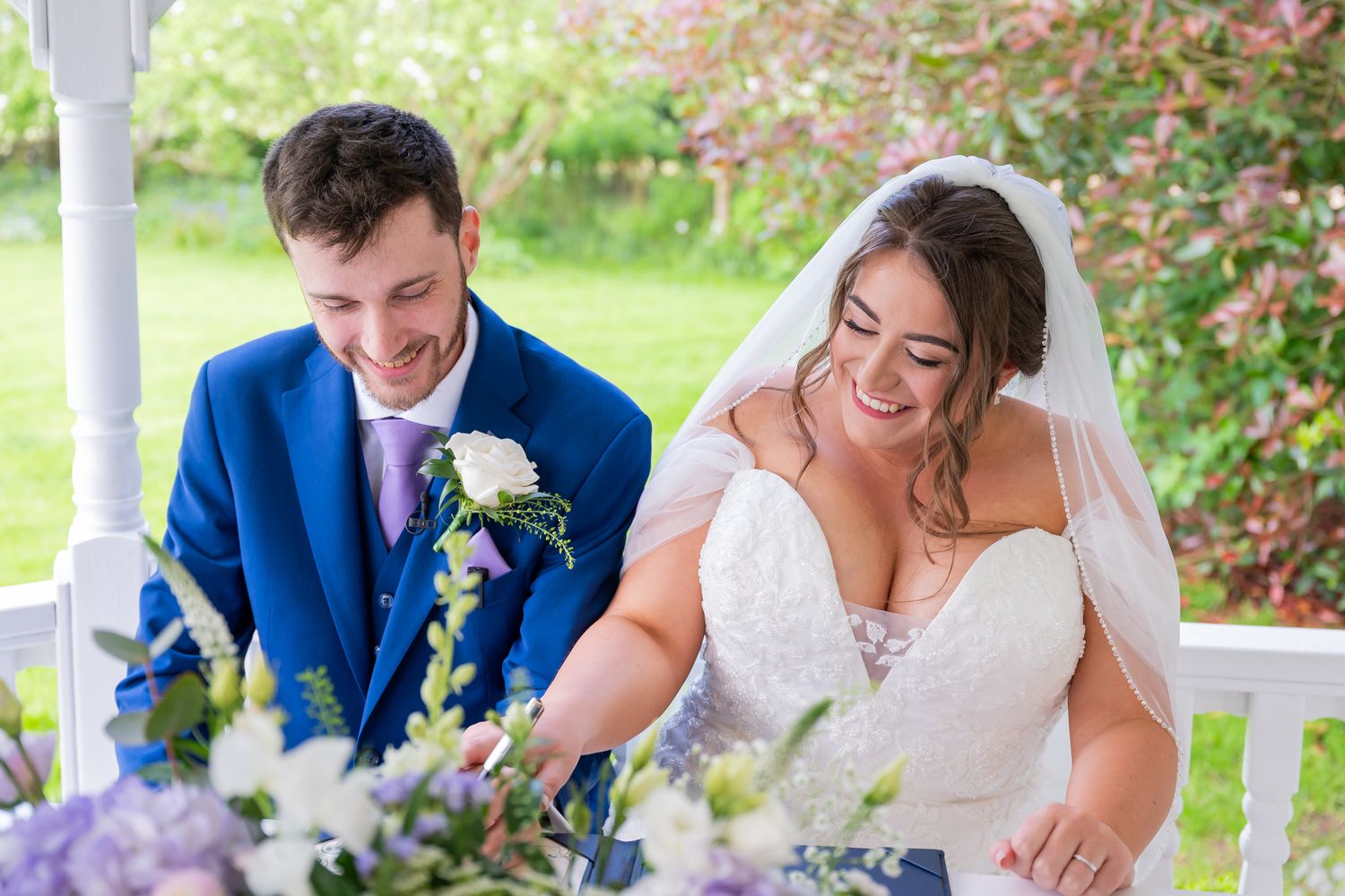 Happy bride and groom signing wedding register, bride in lace gown with veil, groom in blue suit, surrounded by flowers.