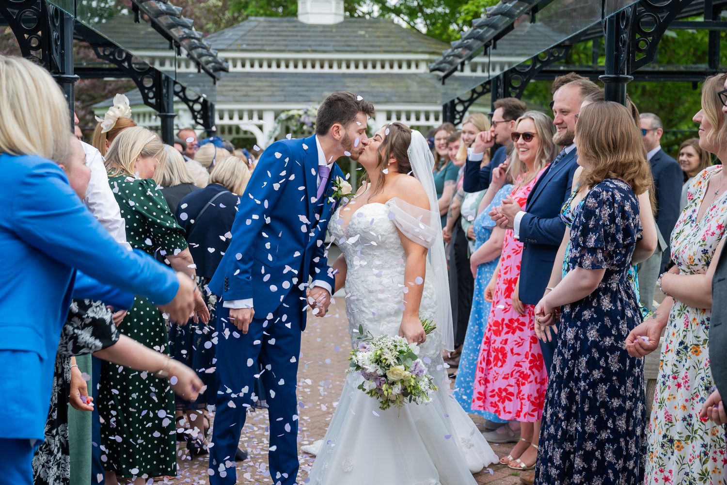 Newlyweds share a kiss during confetti shower surrounded by wedding guests outdoors.