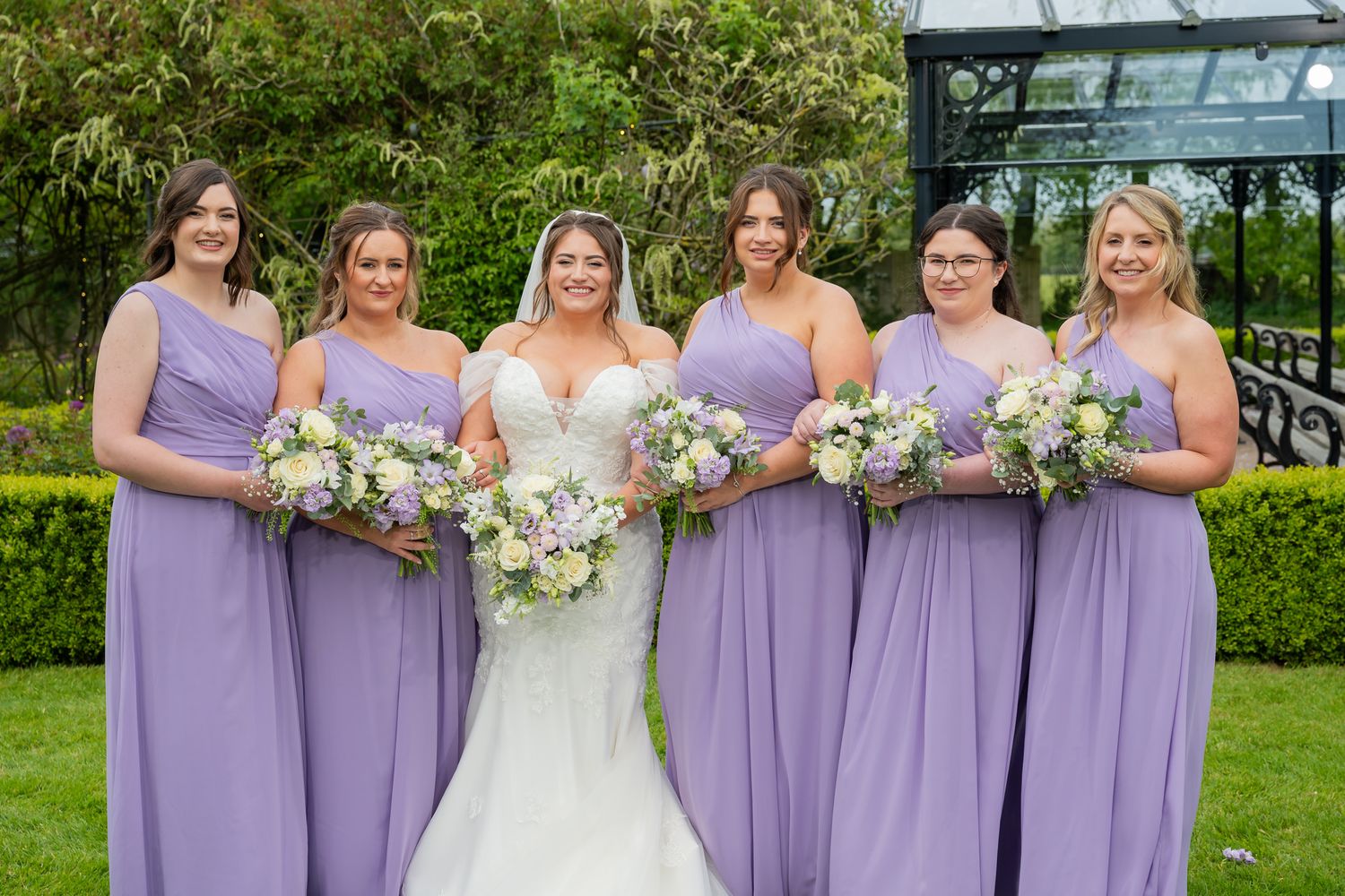 Bride in white gown poses with her bridesmaids in lavender dresses holding bouquets for fun group photos at Old Kent Barn.