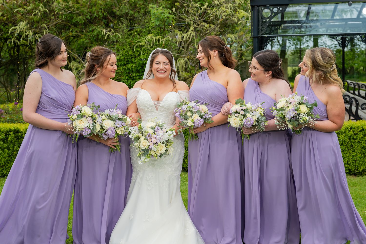 Bride in white lace gown surrounded by five bridesmaids in lavender dresses holding bouquets in a garden setting.