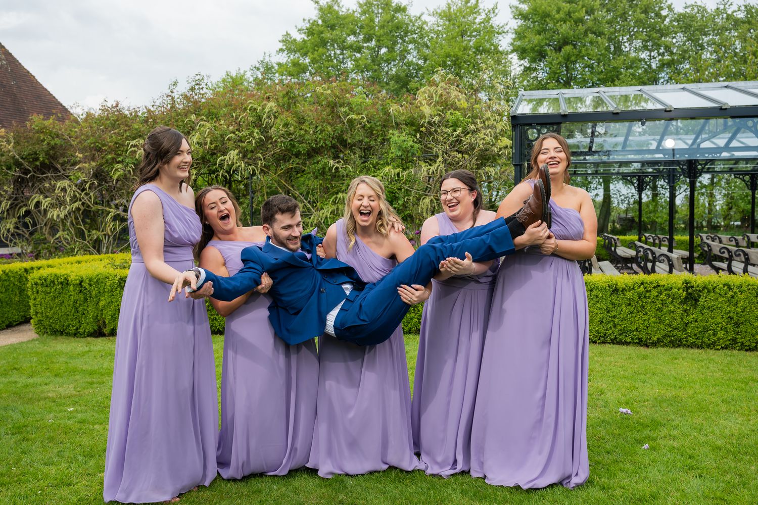 Five bridesmaids in lavender gowns joyfully lift a groom in a navy suit during the group photos at Old Kent Barn.