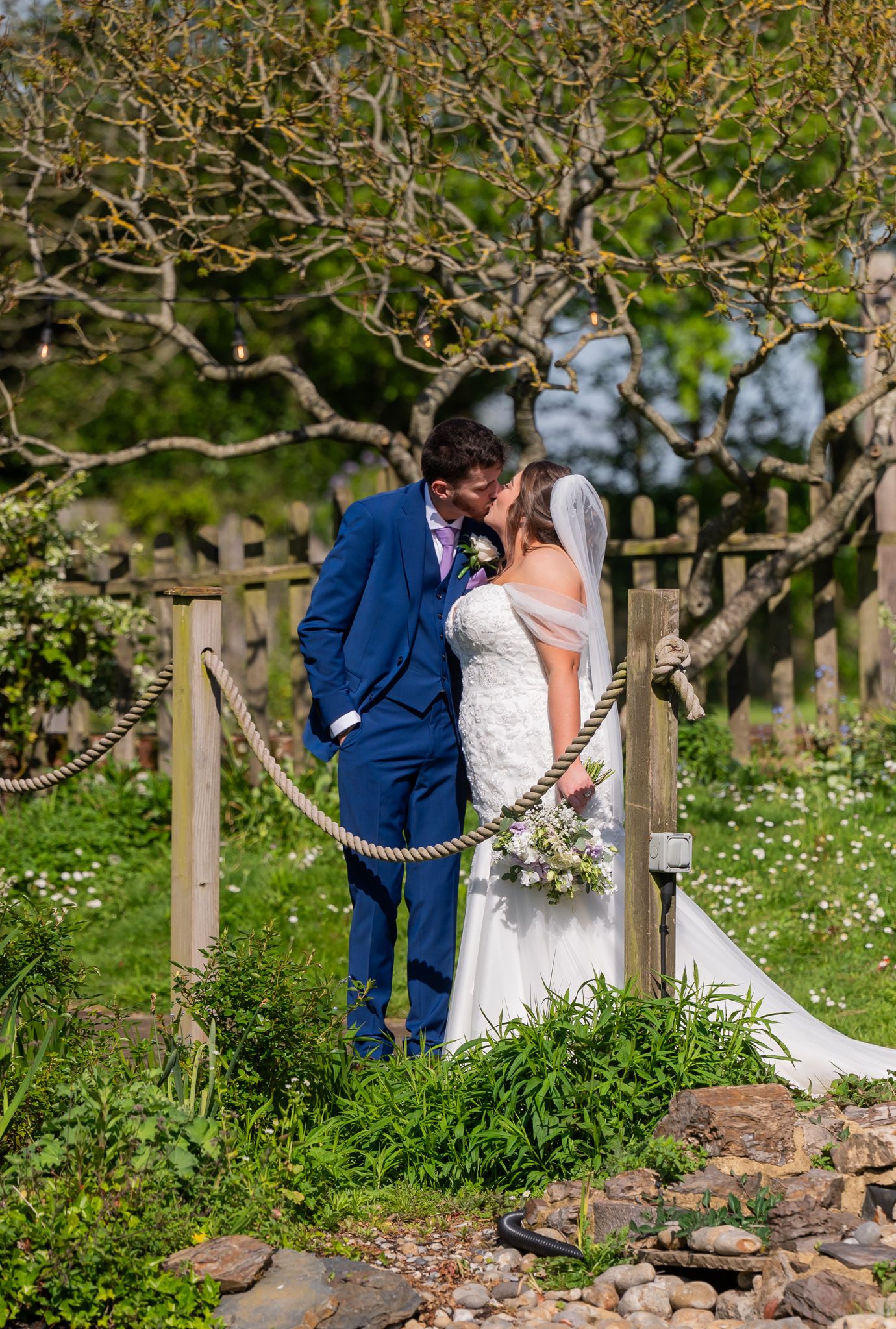 Bride and groom share a romantic kiss in a lush garden setting, with the groom in a blue suit and bride holding a bouquet.