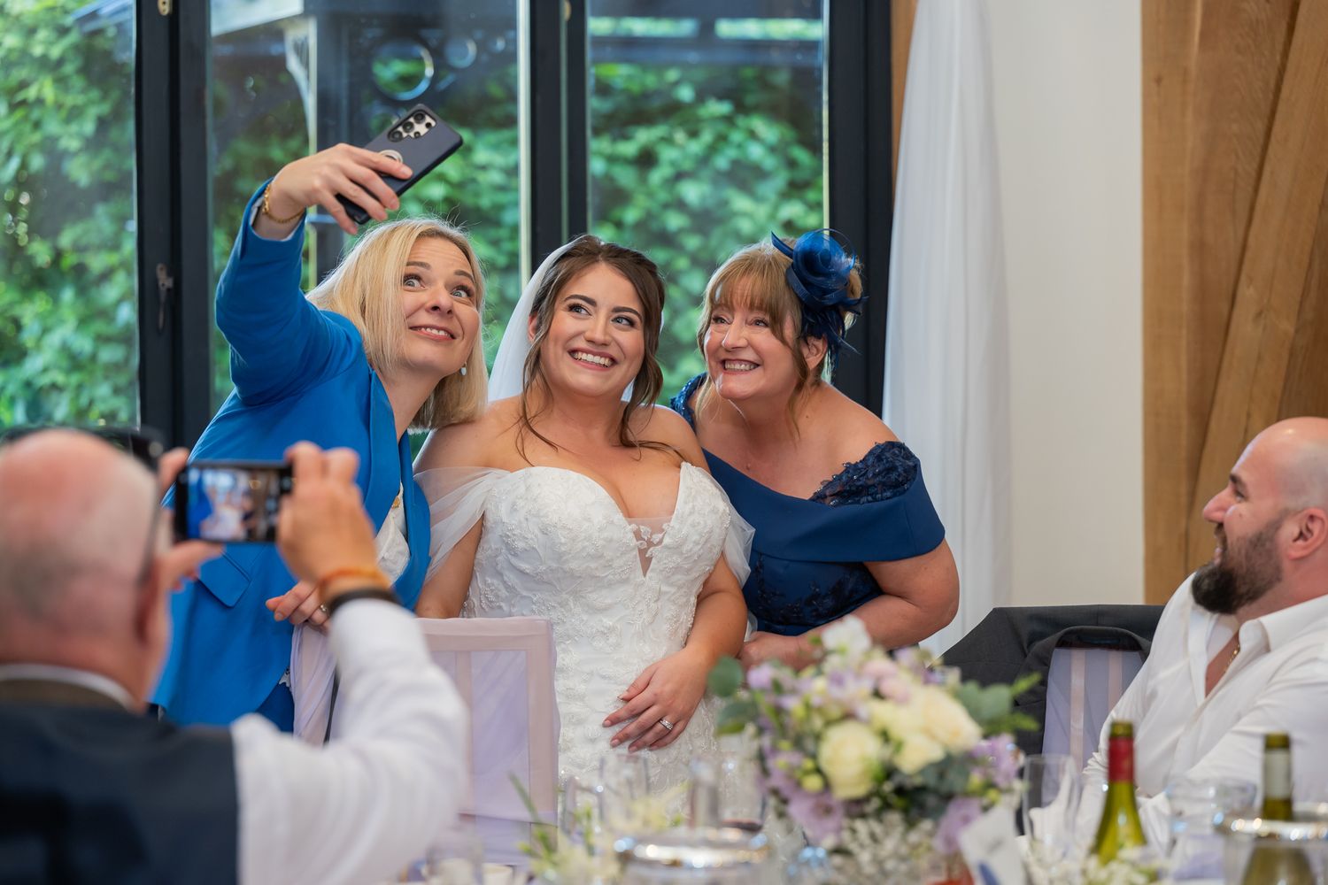 Bride taking a selfie with two guests in blue outfits at a wedding reception dinner table.
