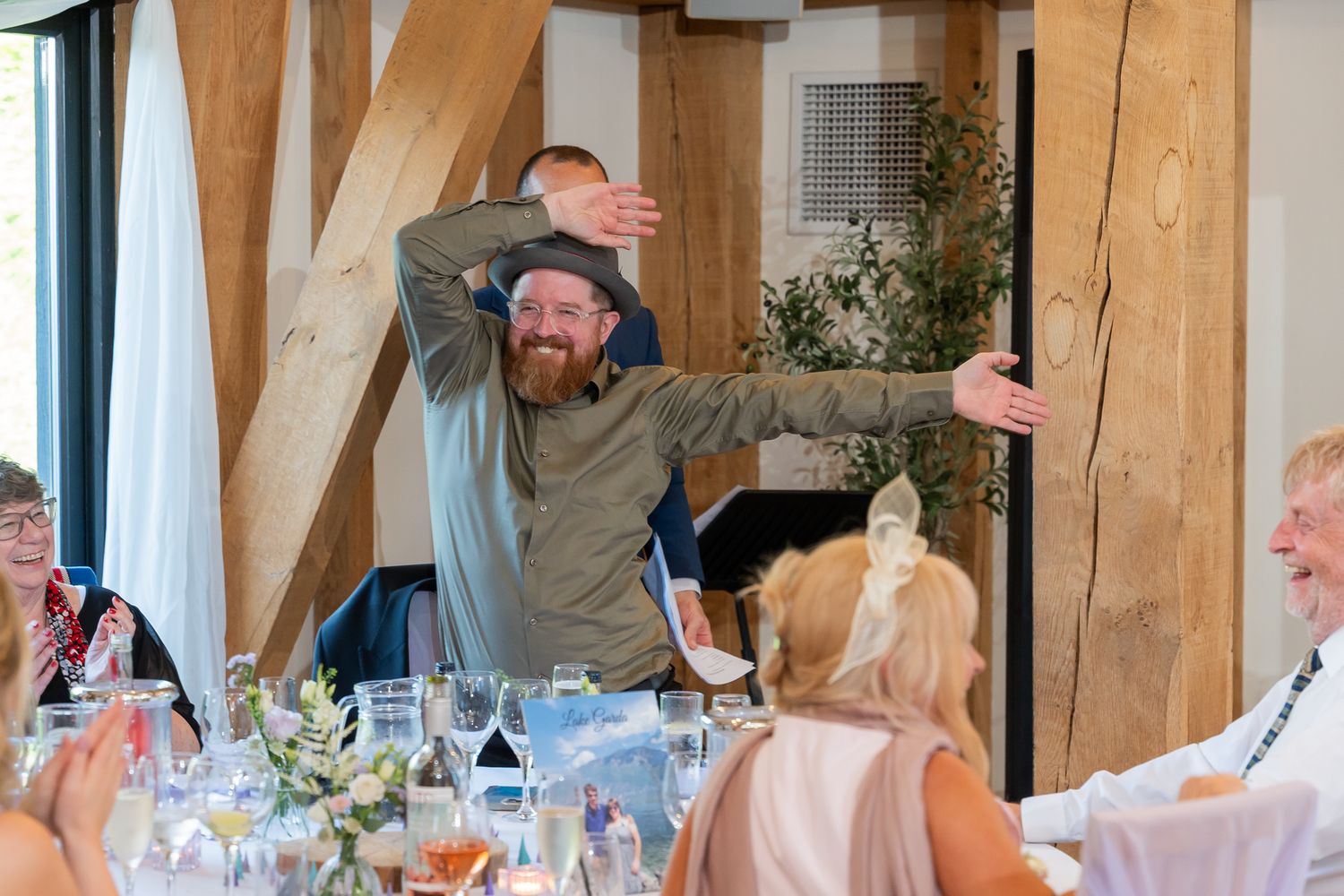Bearded man with glasses raises arms joyfully at a wedding reception dinner table in a rustic barn venue.