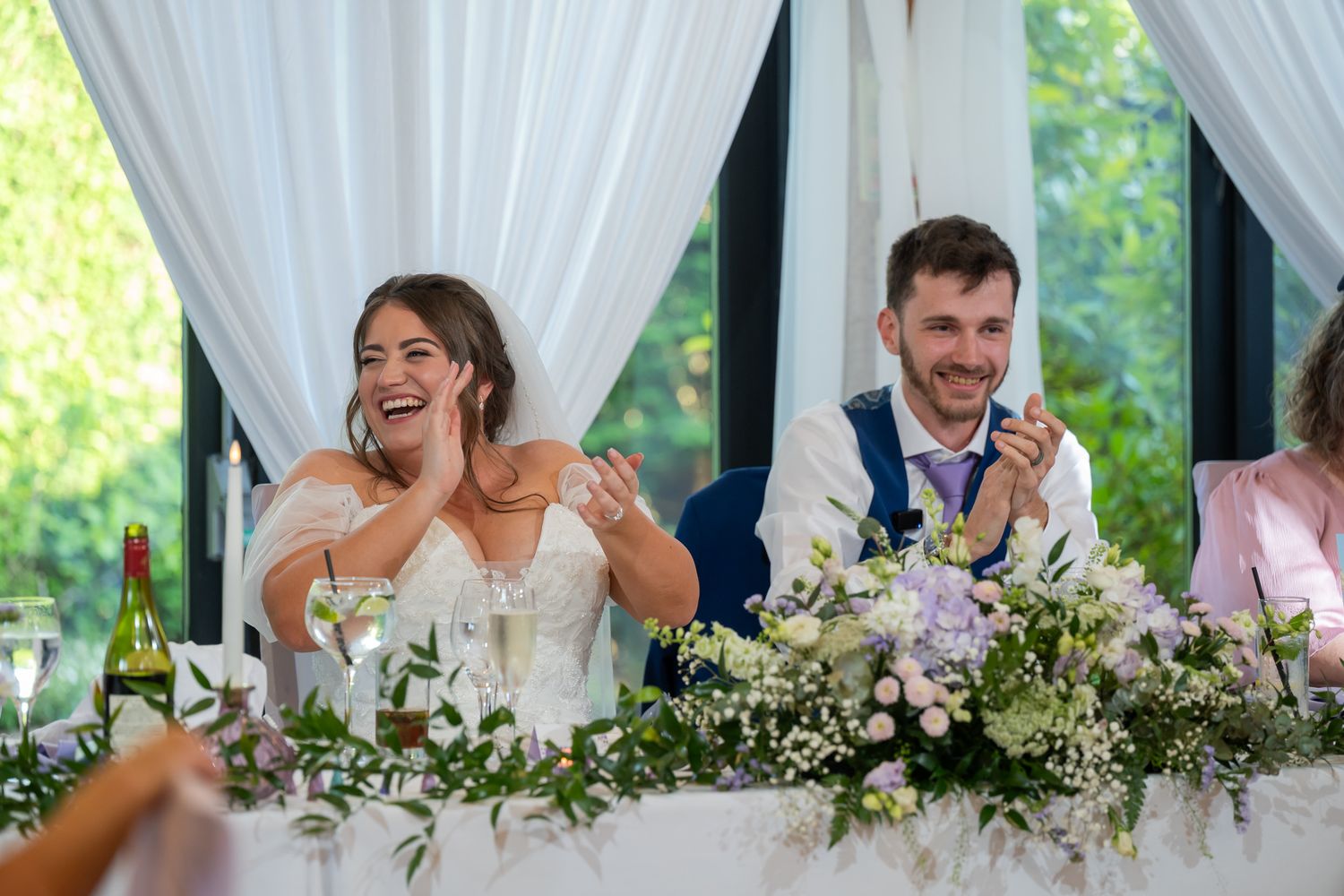 Bride and groom laughing joyfully at wedding reception head table decorated with white flowers and greenery.