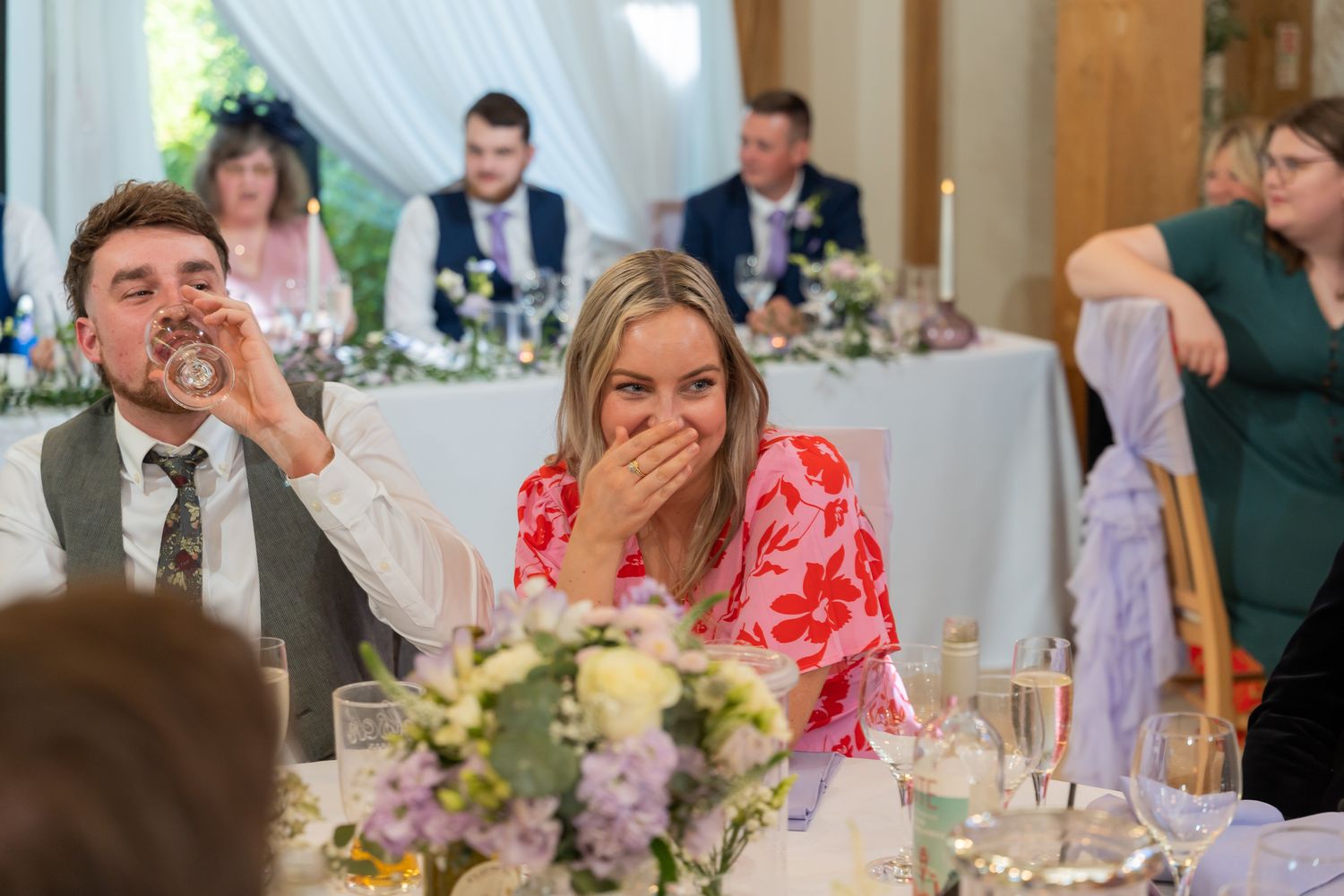 Guests laughing and drinking at a wedding reception dinner table with floral centerpieces.