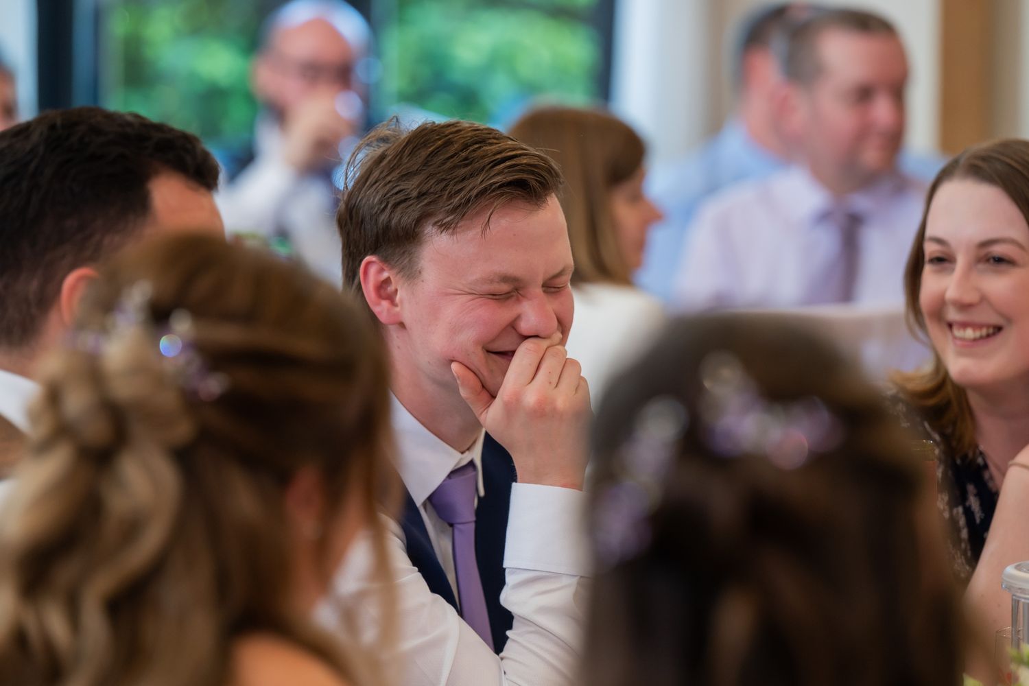 Wedding guest laughing joyfully during reception speeches, wearing suit with purple tie, surrounded by other guests.