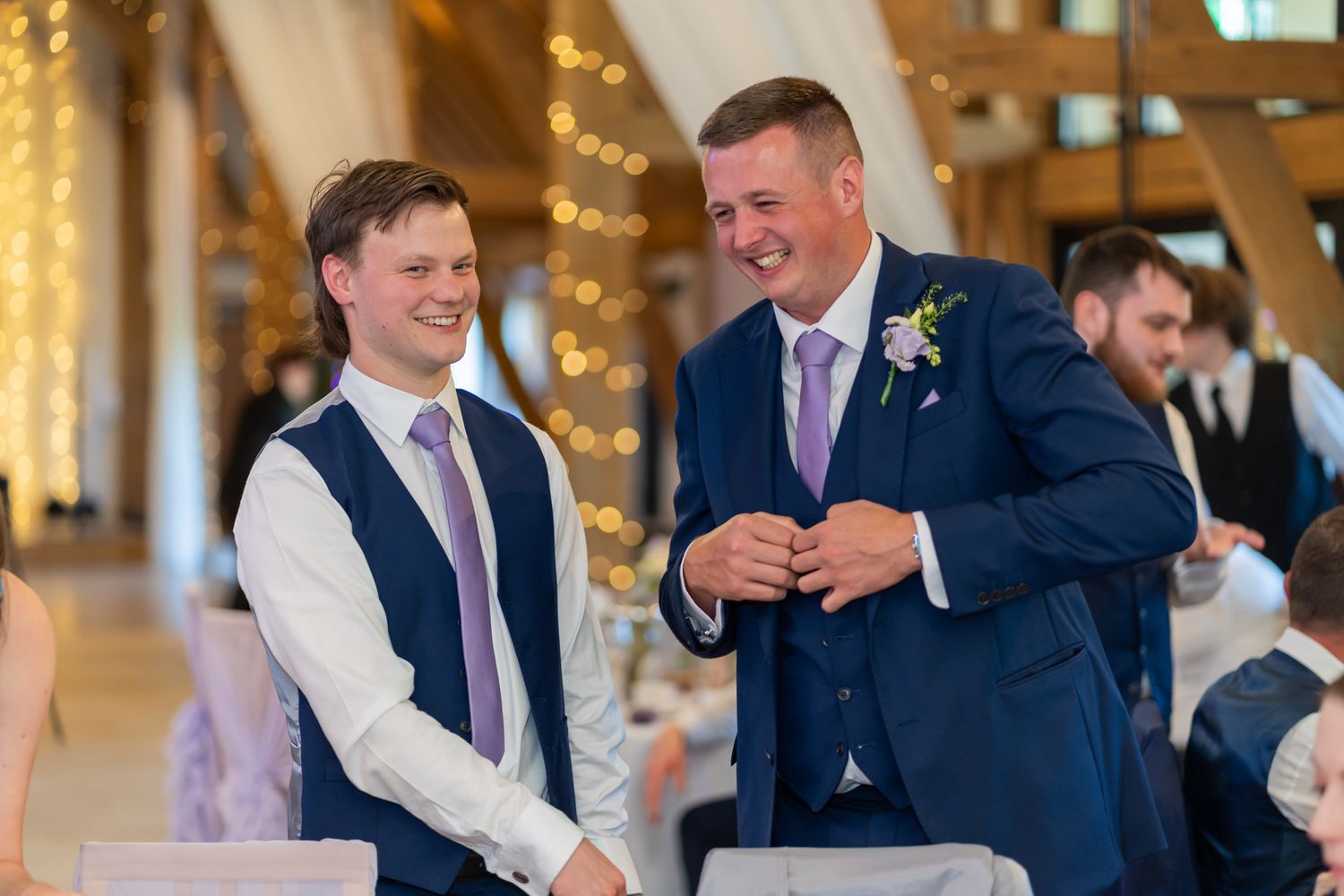 Two men in suits smiling and laughing together at a wedding reception in a decorated barn venue.