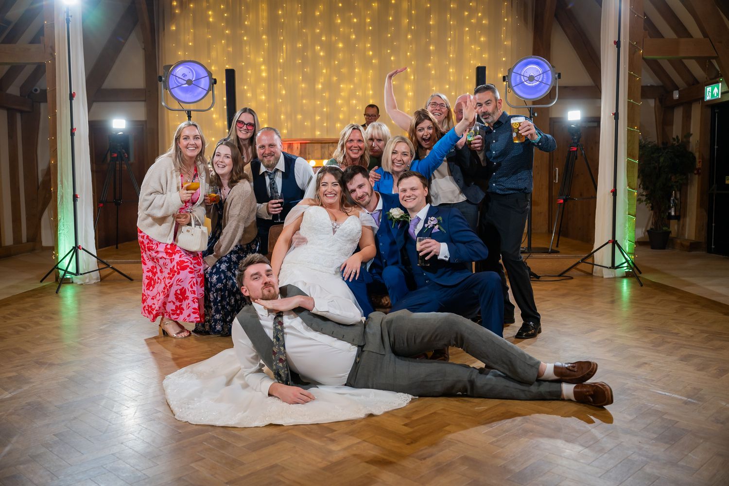 Wedding party celebrating on dance floor with bride and groom surrounded by joyful guests in elegant venue with fairy lights.