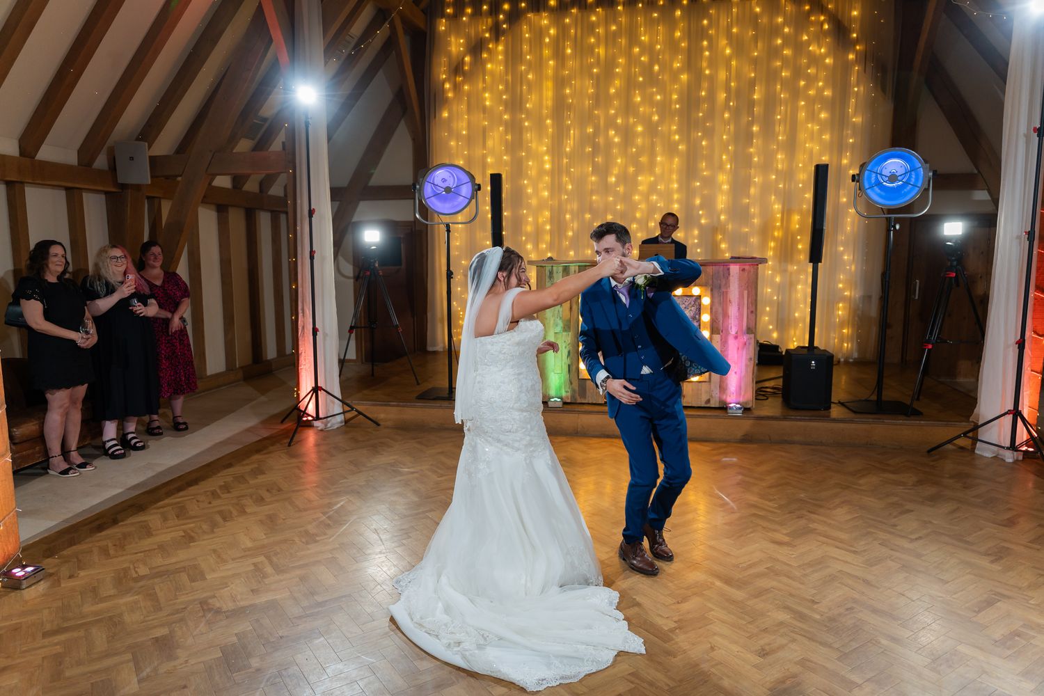 Bride and groom share their first dance in a rustic barn venue with fairy lights and colorful DJ lights.