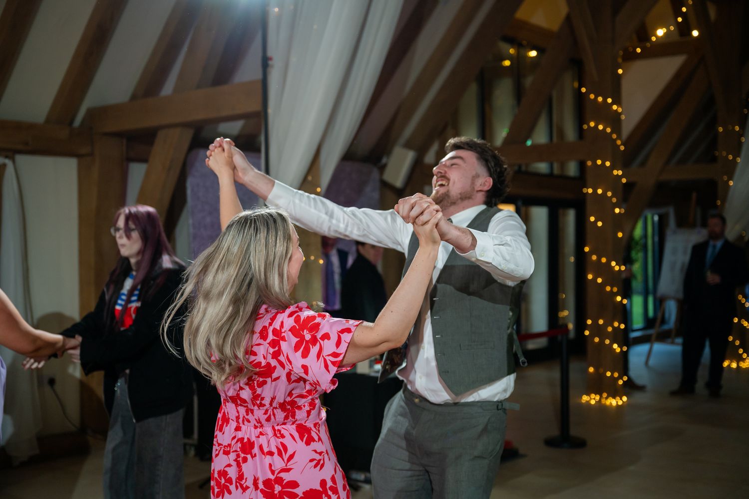 Couple dancing joyfully at a rustic wedding reception with fairy lights and wooden beams in background.