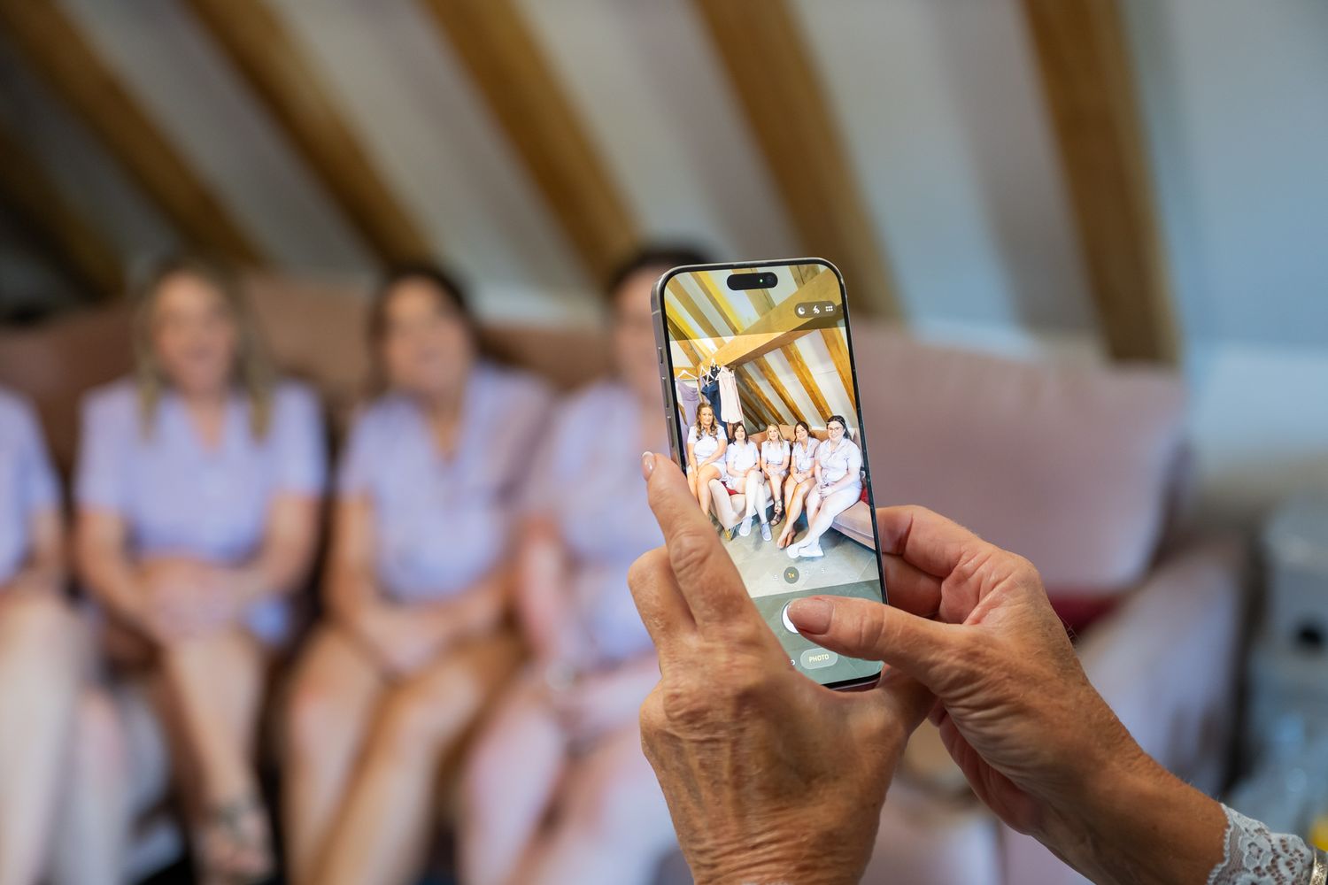 Person photographing bridesmaids in lilac dresses on a smartphone at a wedding preparation.