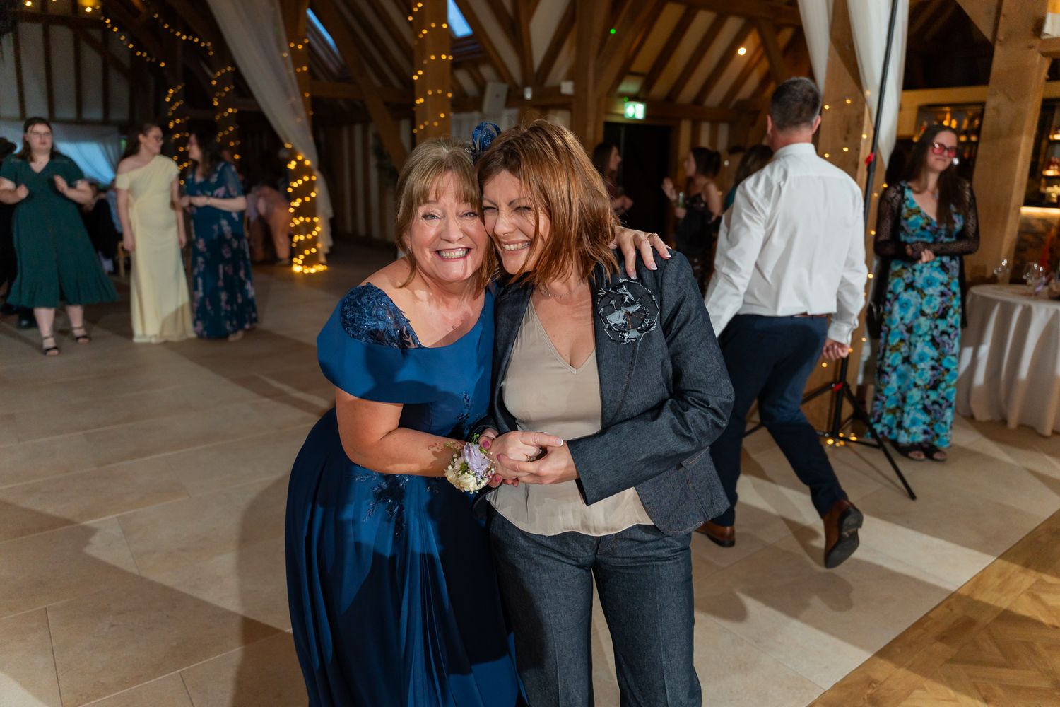 Two women in elegant attire smile and dance together at a wedding reception in a rustic barn venue.