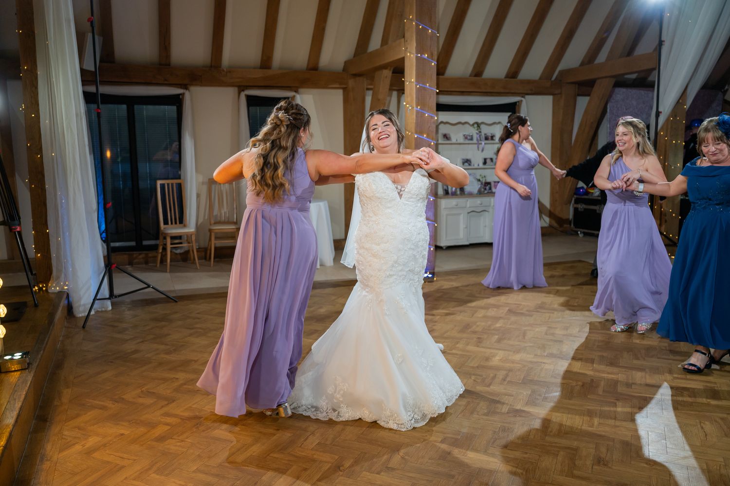 Bride in white mermaid gown dancing with bridesmaids in lavender dresses on wooden dance floor at wedding reception.