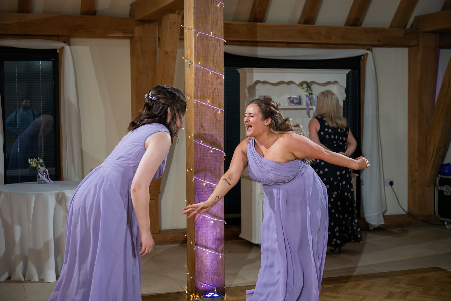Two bridesmaids in lavender gowns dancing and laughing together at a wedding reception venue.