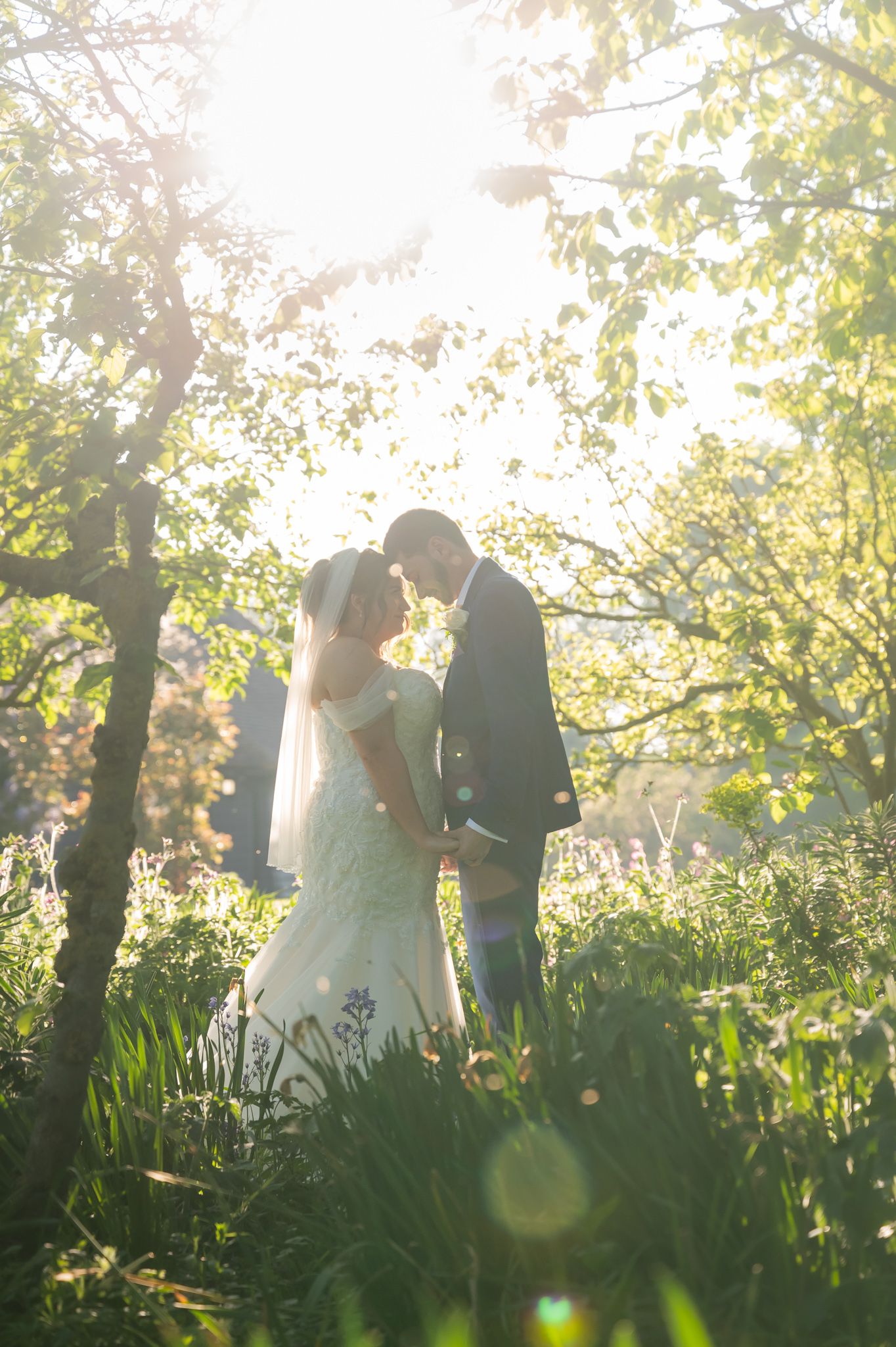 Bride and groom sharing a romantic moment in the sunlit garden, surrounded by lush greenery and soft lens flare at Old Kent Barn near Dover.